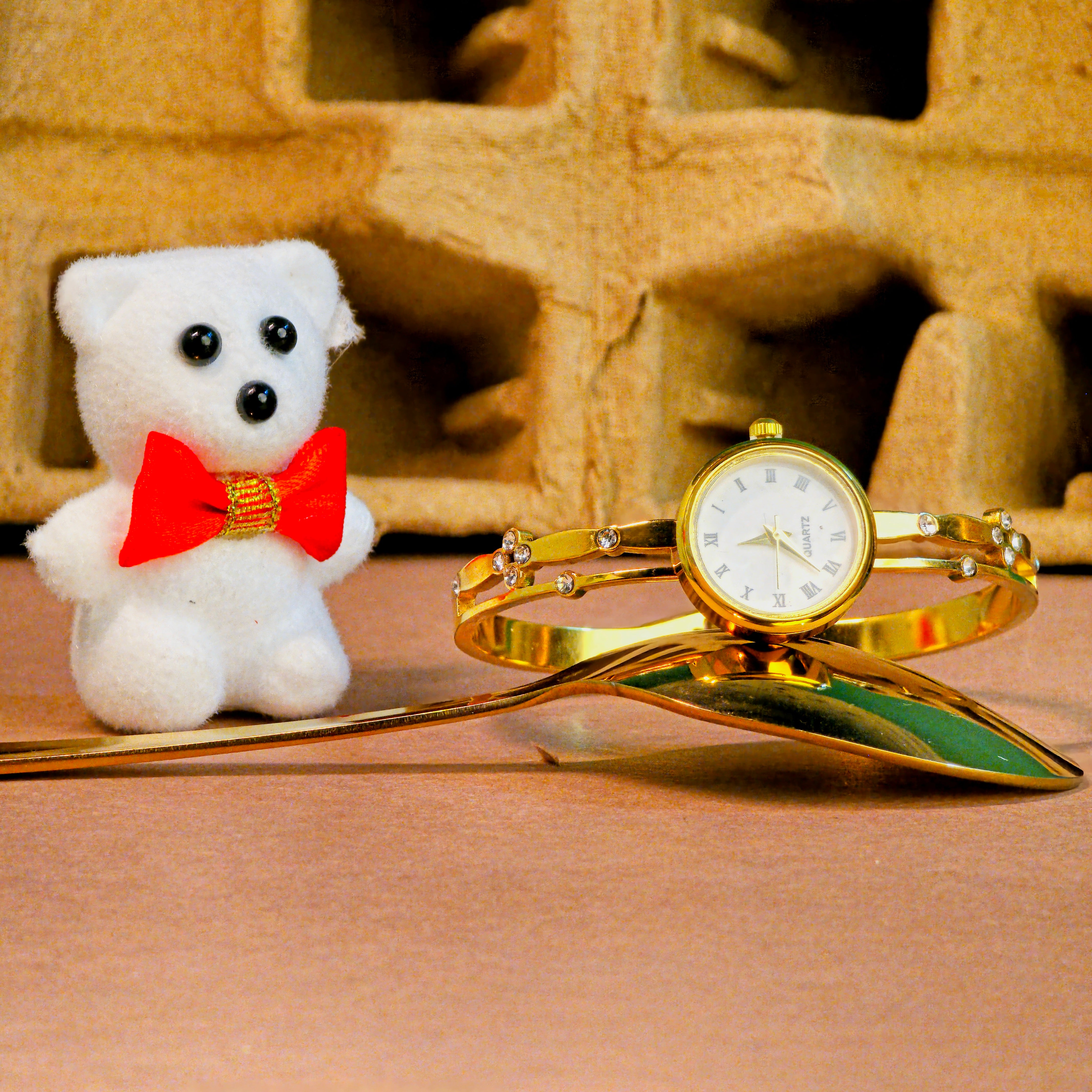 Gold watch with bracelet and small white teddy bear with red bow tie on a textured surface